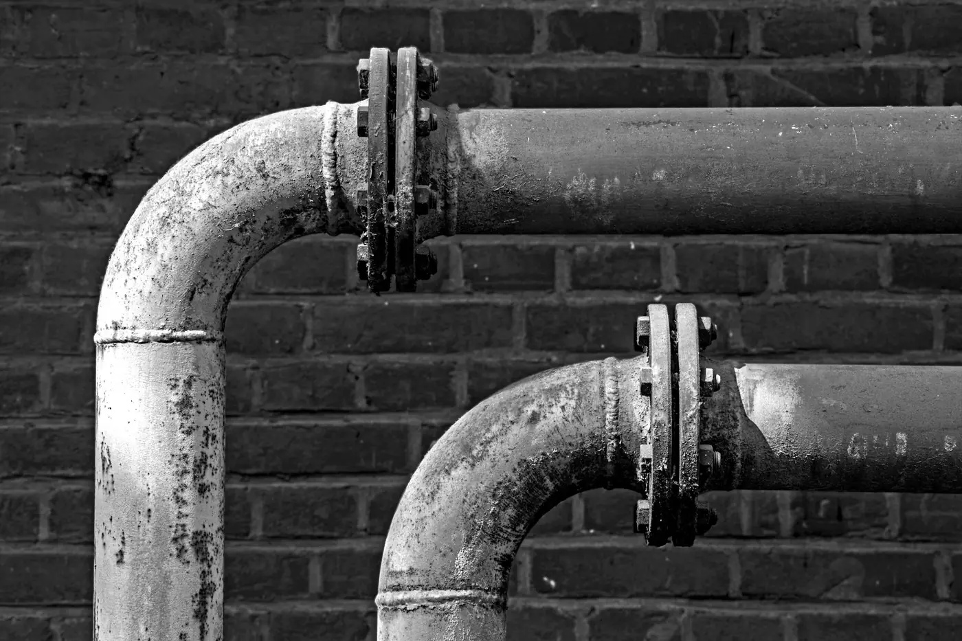 Black and white photo of old flanged metal pipes along a brick wall